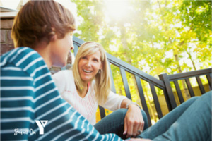 A smiling mother sitting on outdoor deck stairs, having a positive conversation with her teenage son on a brightly lit sunny day, with the YMCA 'Shine On' logo in the bottom left corner.